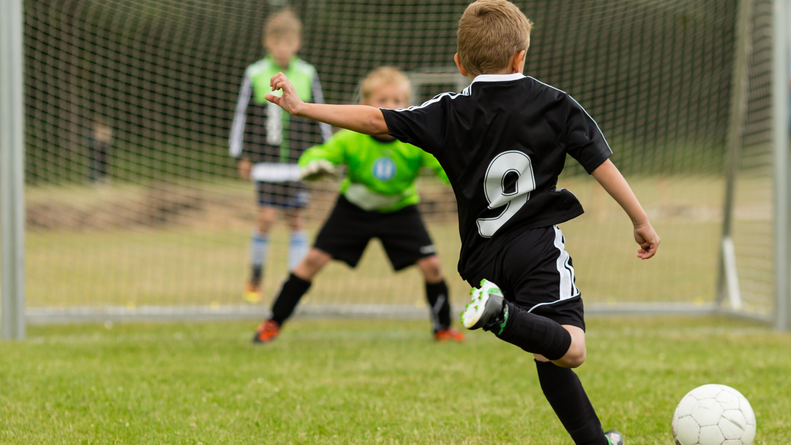 Lapsi potkaisee palloa jalkapallomaalia kohti. Toinen lapsi on taustalla maalissa maalivahtina.