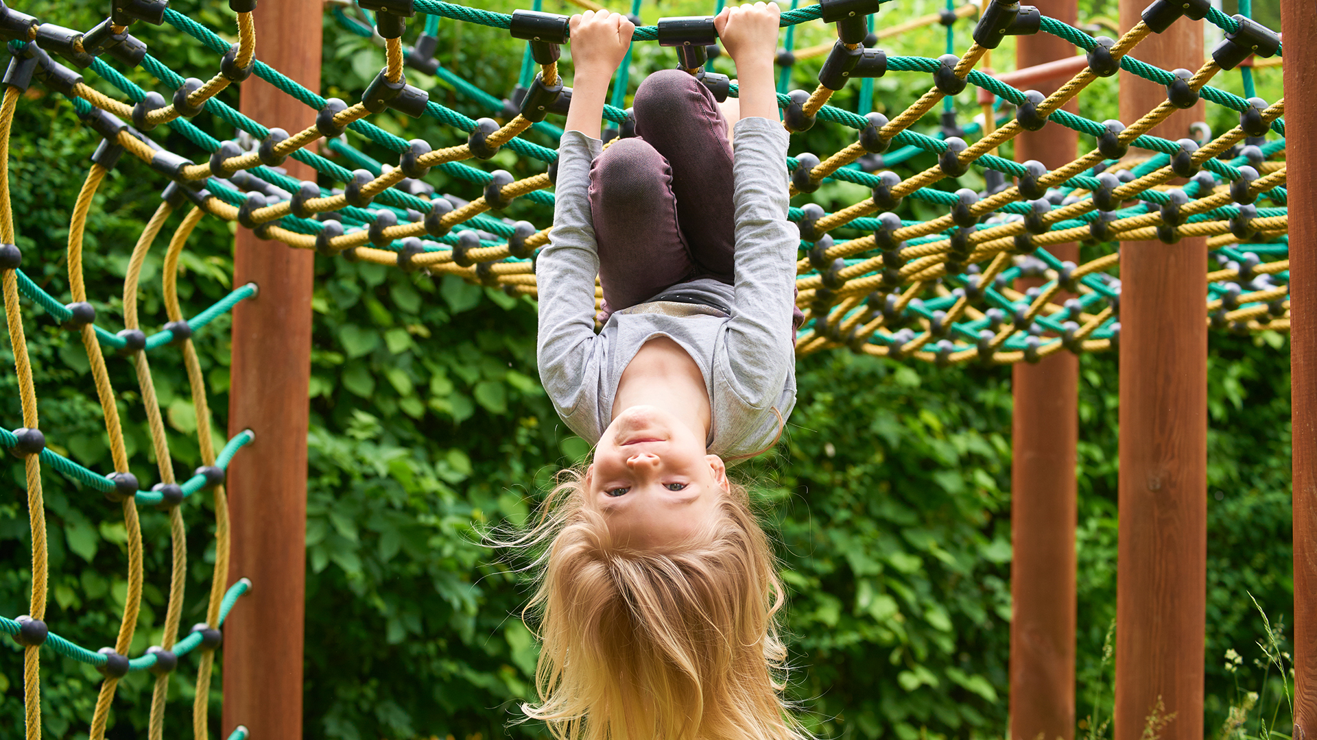 Lapsi roikkuu pää alaspäin kiipeilytelineen verkossa. Taustalla kesäisen vihreää pensasaitaa.