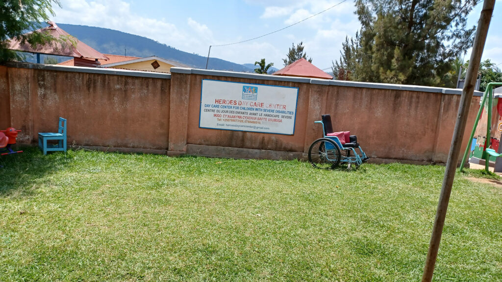 "A picture of a yard with a wall in the background, grass in the foreground, an old-fashioned wheelchair, a small chair, and a bit of a children's swing in the corner."