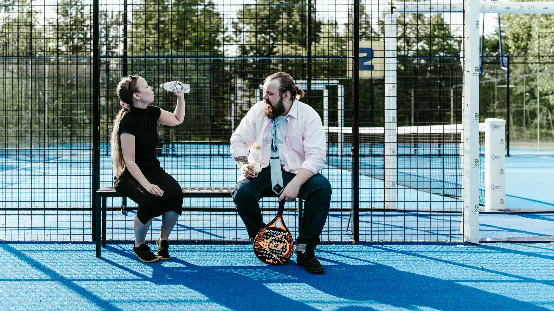 Mies ja nainen istuvat penkillä tenniskentän laidalla vesipullot kädessään. Nainen on juuri hörppäämässä vettä. Miehellä on päällään kauluspaita ja kravatti, naisella tyypillisemmät liikuntavarusteet.