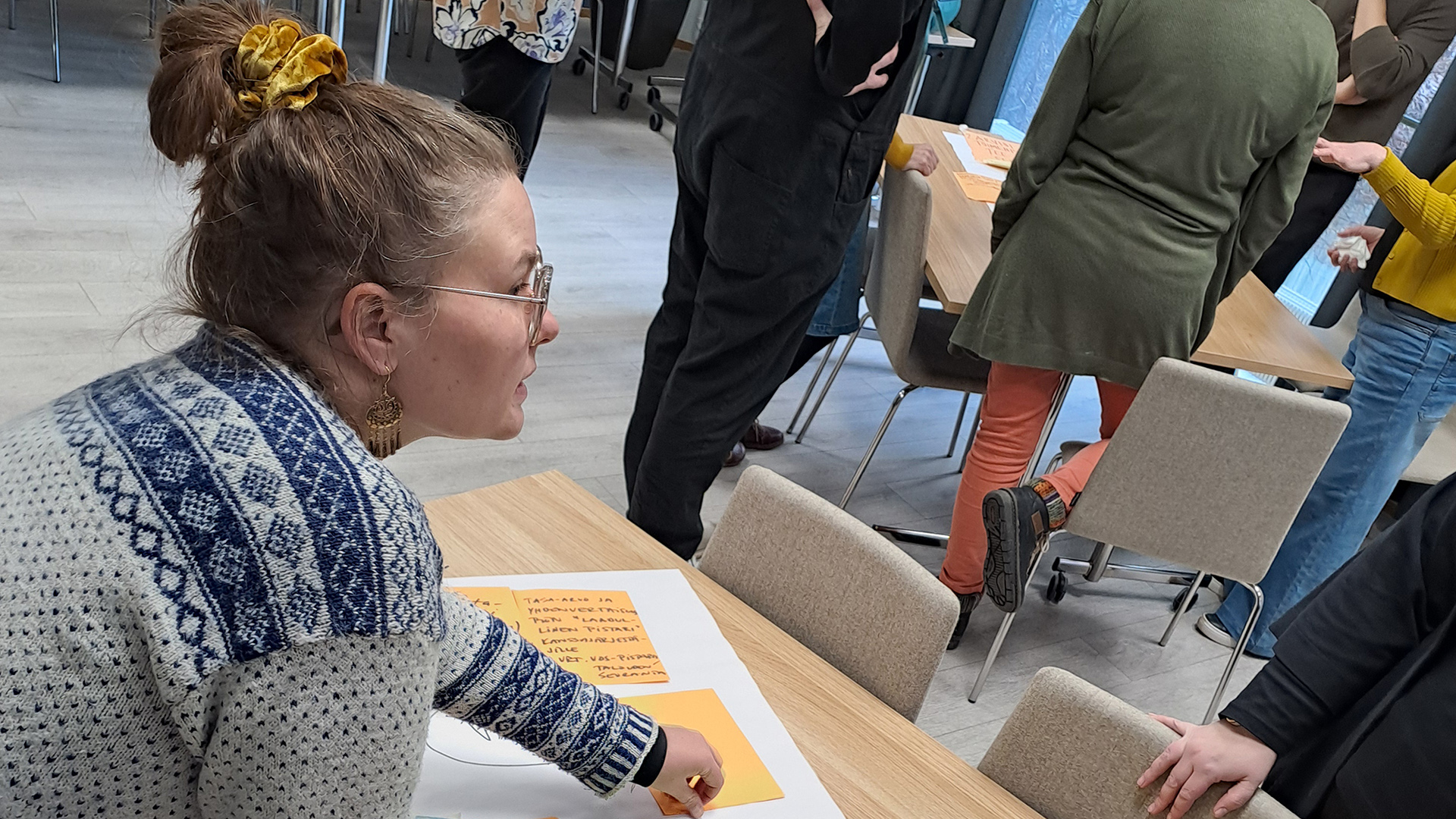 Ihmisiä seisoo työpajatilassa, edustalla yksi henkilö kumartuneena pöydällä olevan paperin ja Postit-lappujen ääreen.