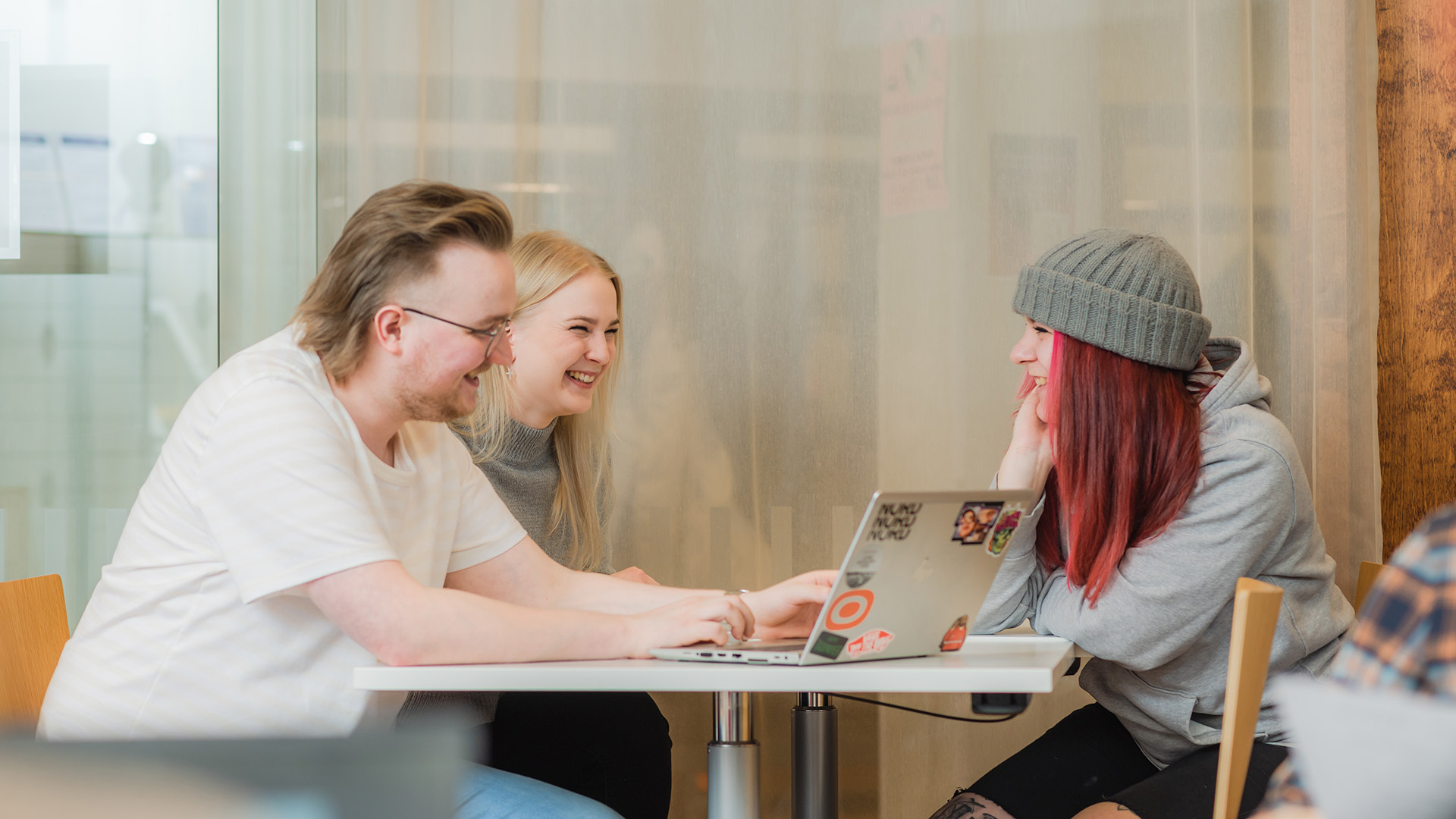 Kolme nuorta istuu pöydän ääressä naureskellen. Yhdellä on läppäri edessään.